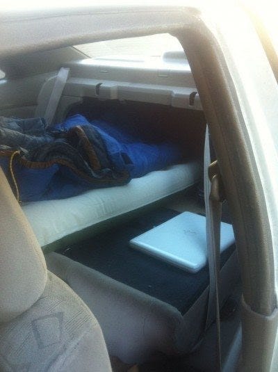 A sleeping bag laid across the back of a car, with a laptop on the rear seat.