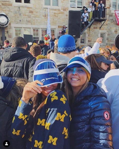 Two people wearing Michigan merchandise at a crowded campus event; one wears a Snackpass-branded hat.